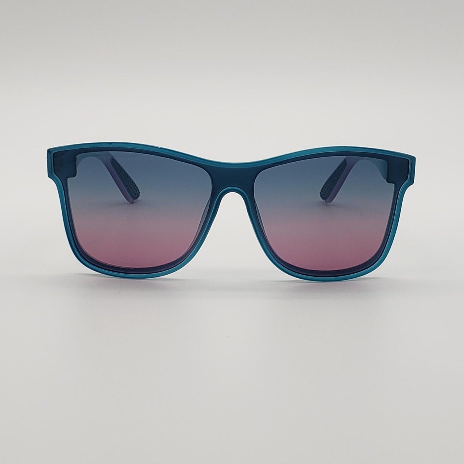 A pair of square sunglasses with a translucent teal-blue frame and gradient lenses that fade from dark blue at the top to pink at the bottom, shown on a neutral light gray background.