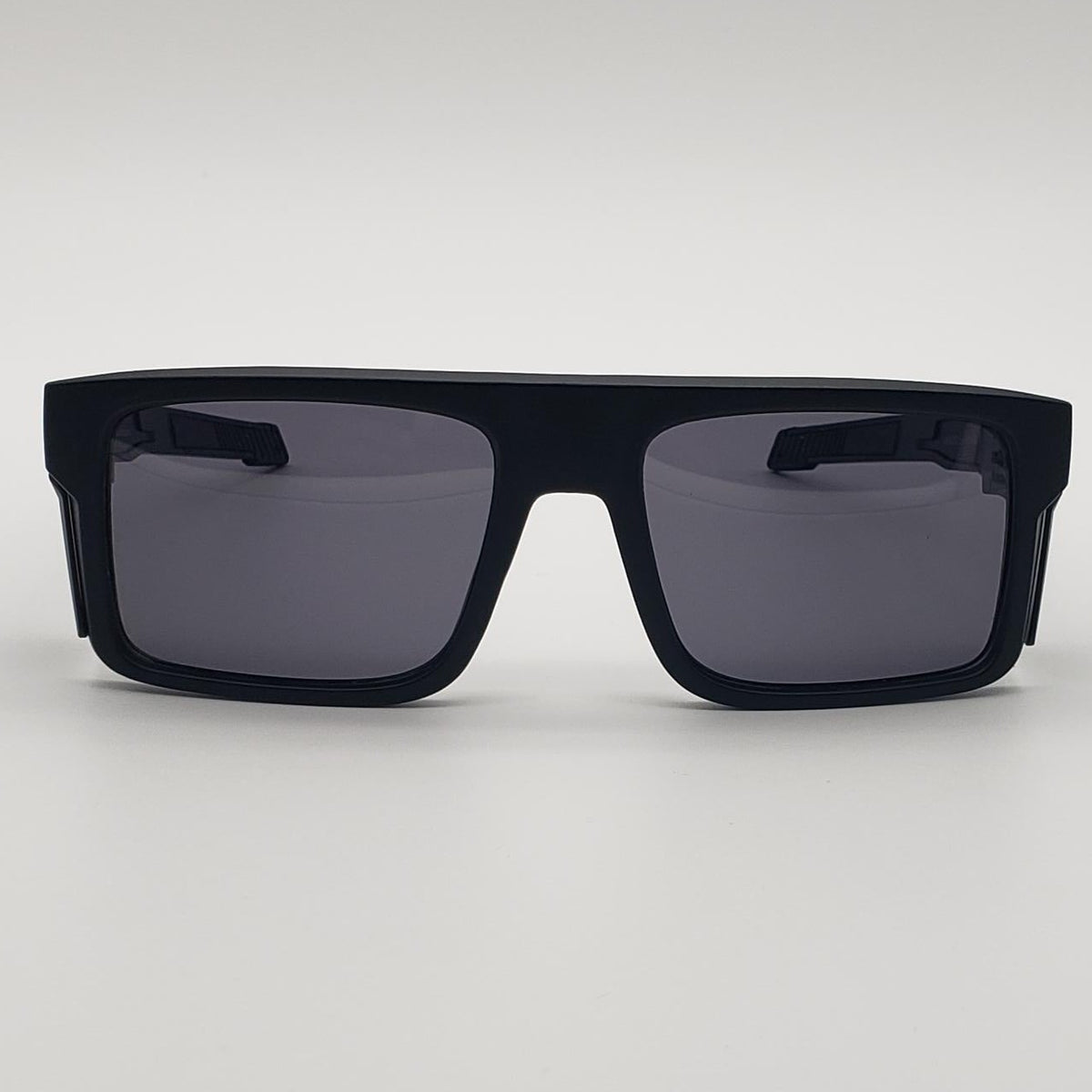 A pair of oversized square sunglasses with a matte black frame and dark gray lenses, photographed against a neutral light gray background.