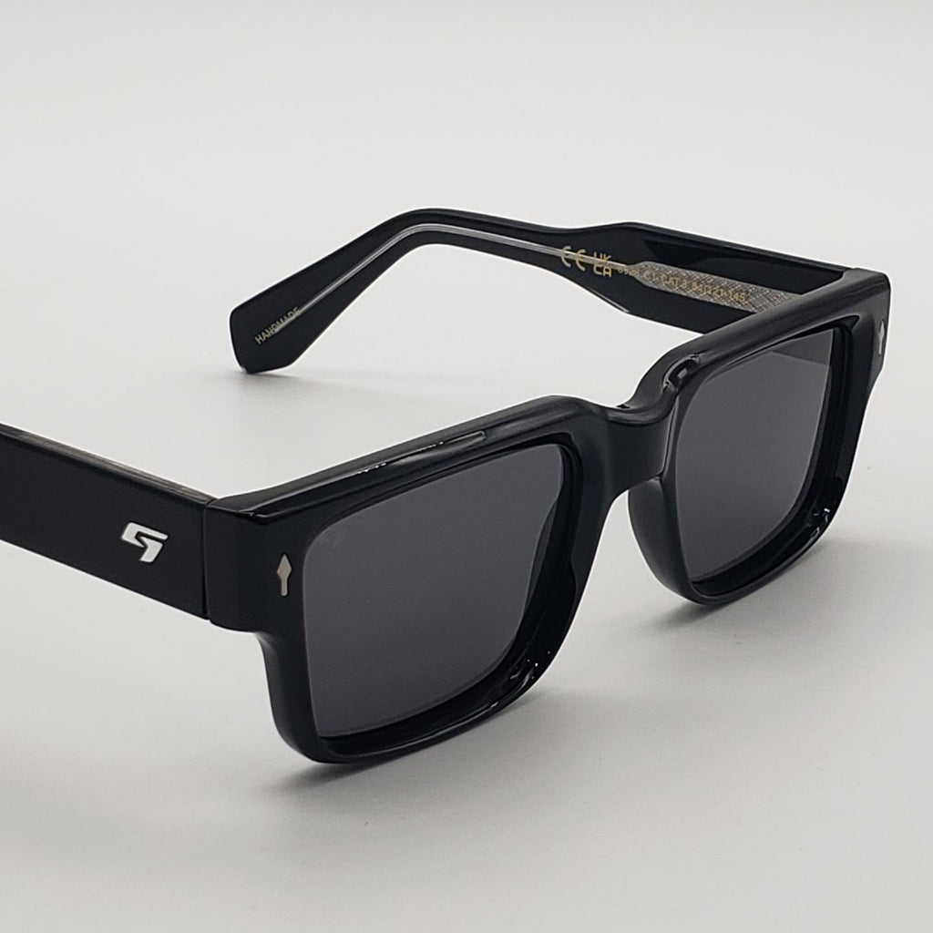 Black square sunglasses with dark lenses and a small white logo on the temple, photographed on a clean light gray background.